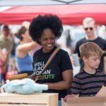 A woman and a boy stand at a market stall with handmade items, surrounded by other people under a canopy. The woman is smiling and holding a clipboard.