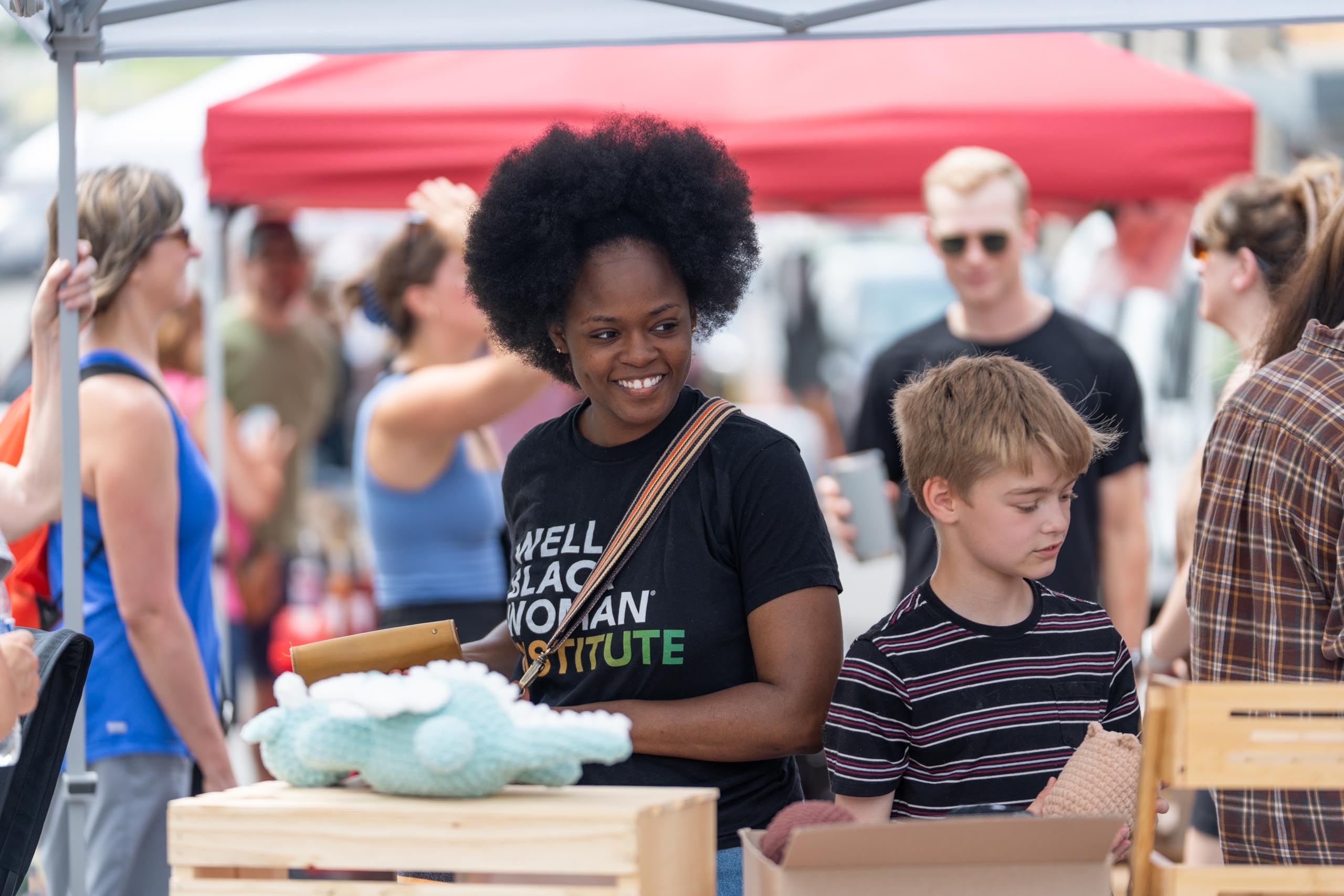 A woman and a boy stand at a market stall with handmade items, surrounded by other people under a canopy. The woman is smiling and holding a clipboard.