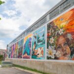A colorful mural on the side of a large building depicts various scenes, including abstract shapes, nature, and a woman’s face with flowers and butterflies.