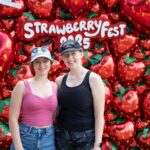 Two people stand in front of a balloon backdrop shaped like strawberries with a sign reading "Strawberry Fest 2023.