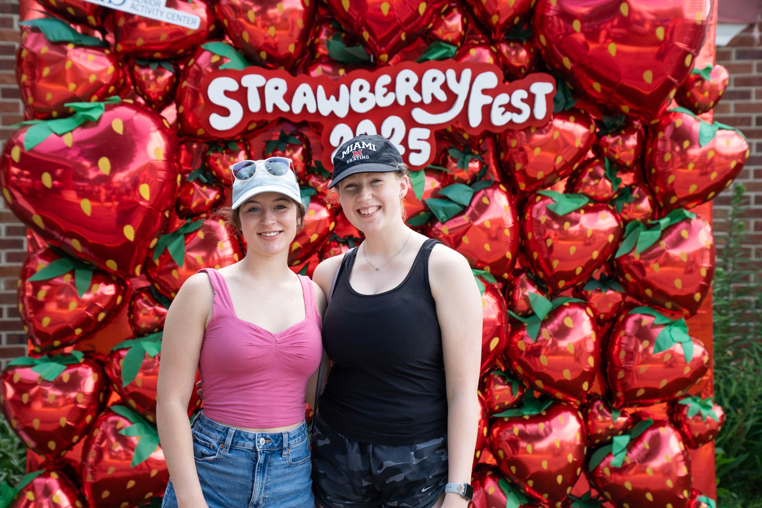 Two people stand in front of a balloon backdrop shaped like strawberries with a sign reading "Strawberry Fest 2023.