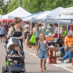 A woman pushes a stroller with a child while another child walks beside her, eating ice cream, at an outdoor market with white tents and people in the background.