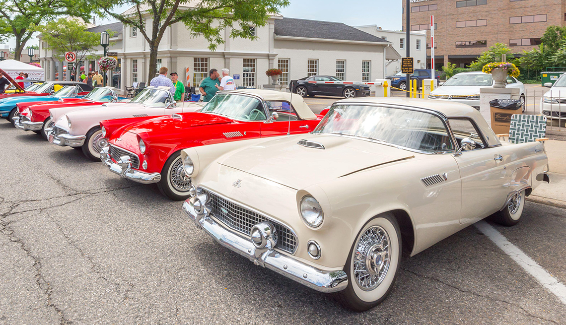 A row of classic vintage cars, including white and red convertibles, is displayed at a car show on a city street with people walking nearby.