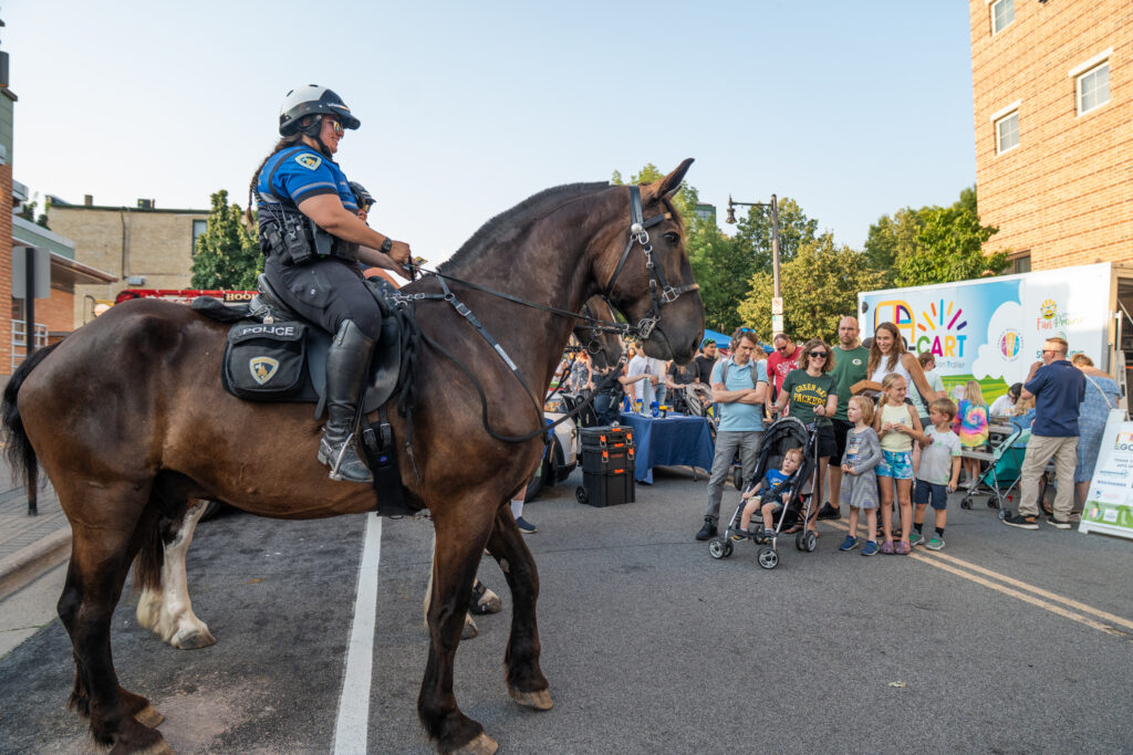 A police officer on a horse interacts with a group of adults and children at an outdoor community event on a city street.