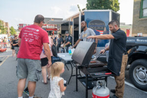 People gather around a grill at an outdoor community event, with police officers and volunteers serving food near a Sun Prairie Police trailer.