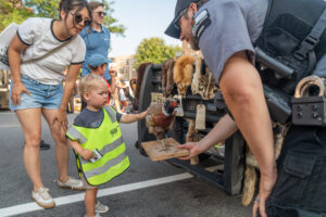 A police officer shows a taxidermy bird to a young child wearing a safety vest at an outdoor event, while adults observe nearby.
