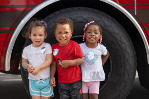 Three young children stand smiling in front of a large vehicle tire, wearing casual summer clothes and sticker badges.