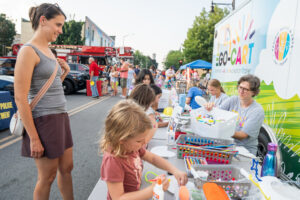 A group of children and adults participate in a craft activity at a table set up on a blocked-off city street during an outdoor event.