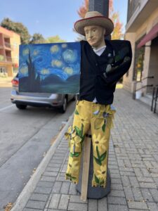 A scarecrow dressed as an artist holds a painting resembling Van Gogh's "Starry Night" while standing on a city sidewalk next to a parked car.