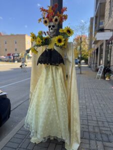 A skeleton dressed in a yellow polka-dot gown with sunflowers and autumn leaves on its head stands on a city sidewalk.