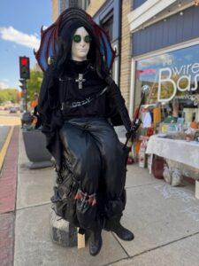 A mannequin dressed in all black with goggles, a cross necklace, and bat-like wings sits on a sidewalk outside a vintage shop.