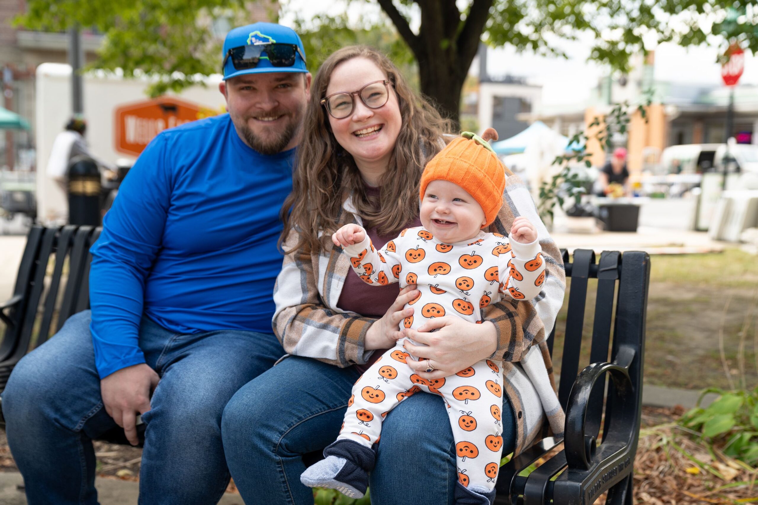 A man, a woman, and a baby in a pumpkin-themed outfit sit on a park bench, smiling at the camera with trees and buildings in the background.