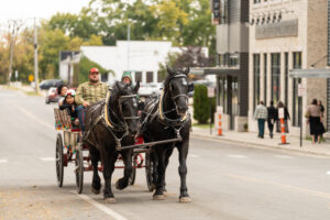 A man and children ride a horse-drawn carriage down a city street; pedestrians walk on the sidewalk in the background.