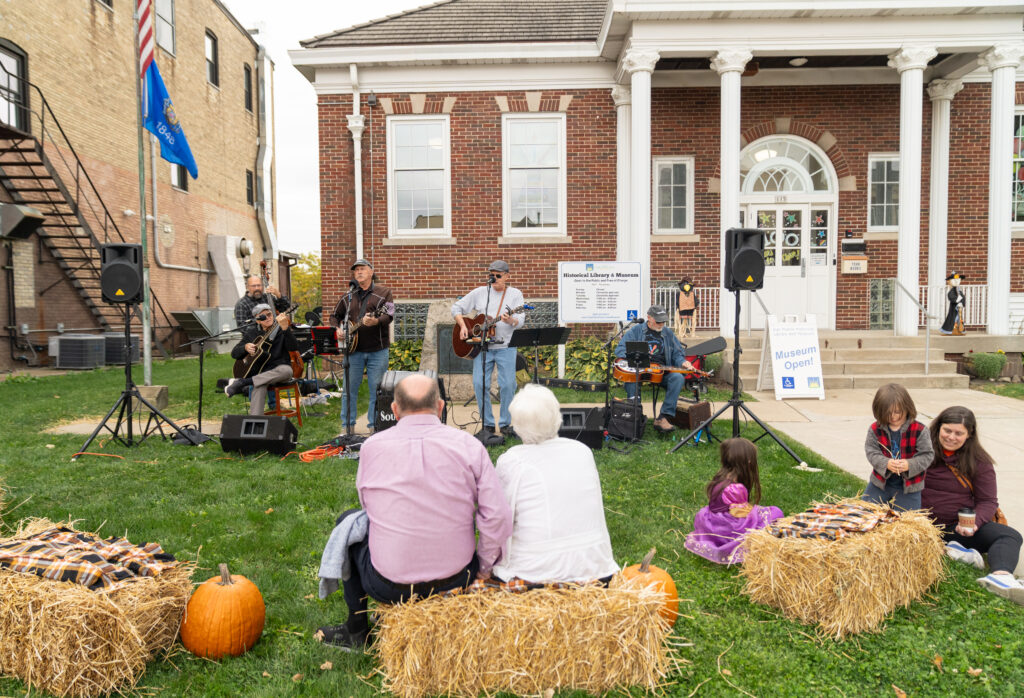 A small band performs outdoors in front of a brick building as people sit on hay bales and listen; pumpkins and autumn decorations are on the grass.