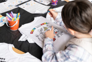 A child colors a Halloween-themed T-shirt with markers at a table covered in art supplies and printed instructions.