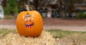 A pumpkin decorated with a cartoon monster face sticker sits on a bale of straw outdoors.