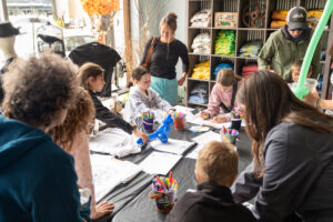 Children and adults gather around a table covered with art supplies, coloring and drawing on white T-shirts in a store with shelves of folded clothes in the background.