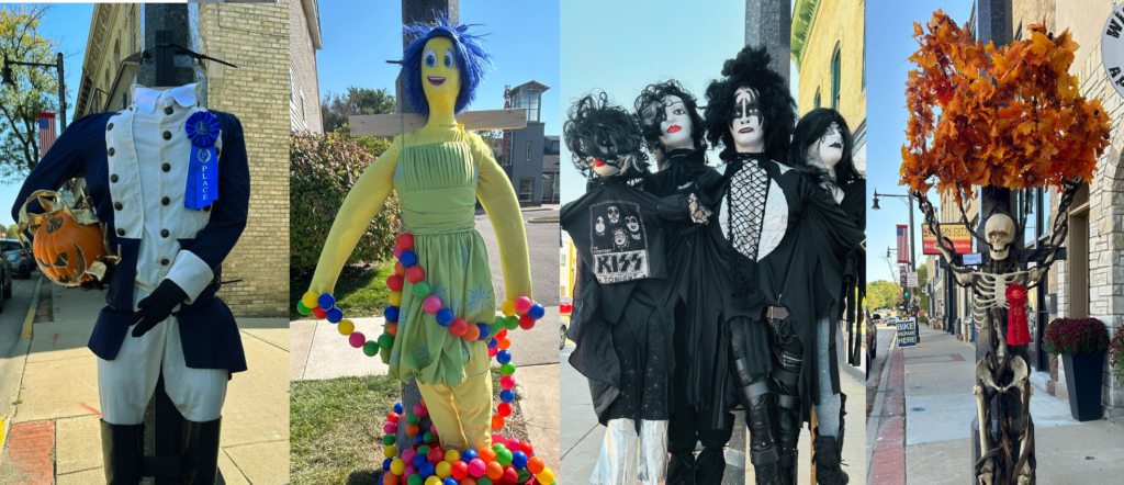 Four scarecrows stand on a sidewalk: a colonial soldier with a blue ribbon, a yellow figure in a green dress, three figures dressed as KISS band members, and a skeleton holding orange leaves.