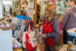 A woman in a red jacket smiles while holding her wallet and shopping in a store filled with bags, soap, and various gift items. Another person browses nearby.