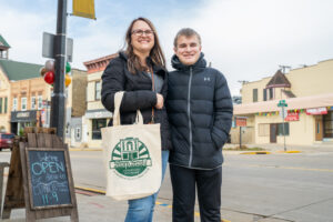 A woman and a teenage boy stand on a sidewalk in a small town. The woman holds a "Shop Small" tote bag. Shops and a sign listing lunch hours are visible in the background.