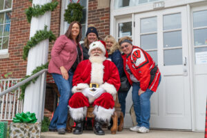 Four women pose and smile around a man dressed as Santa Claus seated outside a brick building decorated with garland and presents.