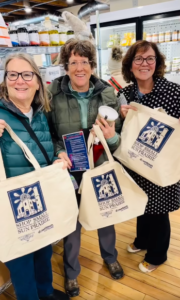 Three women stand indoors holding beige "Shop Small Sun Prairie" tote bags and smiling, with shelves of products and a refrigerator in the background.