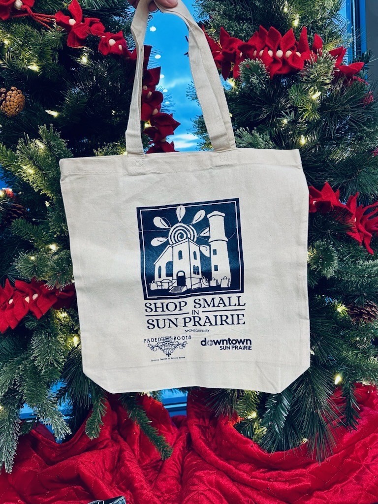A hand holds a tote bag in front of decorated Christmas trees. The bag reads "Shop Small in Sun Prairie" with a graphic of buildings and "downtown Sun Prairie" text.