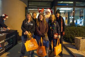 A group of six people, some holding orange shopping bags and wearing festive accessories, pose and smile for a photo at an outdoor event in the evening.