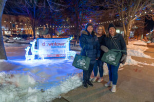 Three women in winter coats and hats stand on a snowy sidewalk at night in downtown Sun Prairie, posing near a lit holiday display and holding tote bags.