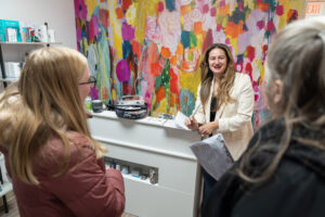 A woman stands behind a reception desk, smiling and talking to two other women in a colorful, modern office or clinic.