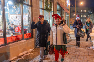 Two people in winter clothing, one dressed festively, walk on a snowy sidewalk at night, smiling and waving, with others and decorated shop windows in the background.
