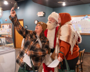 Two women take a selfie with a person dressed as Santa Claus inside a room decorated with winter holiday items.