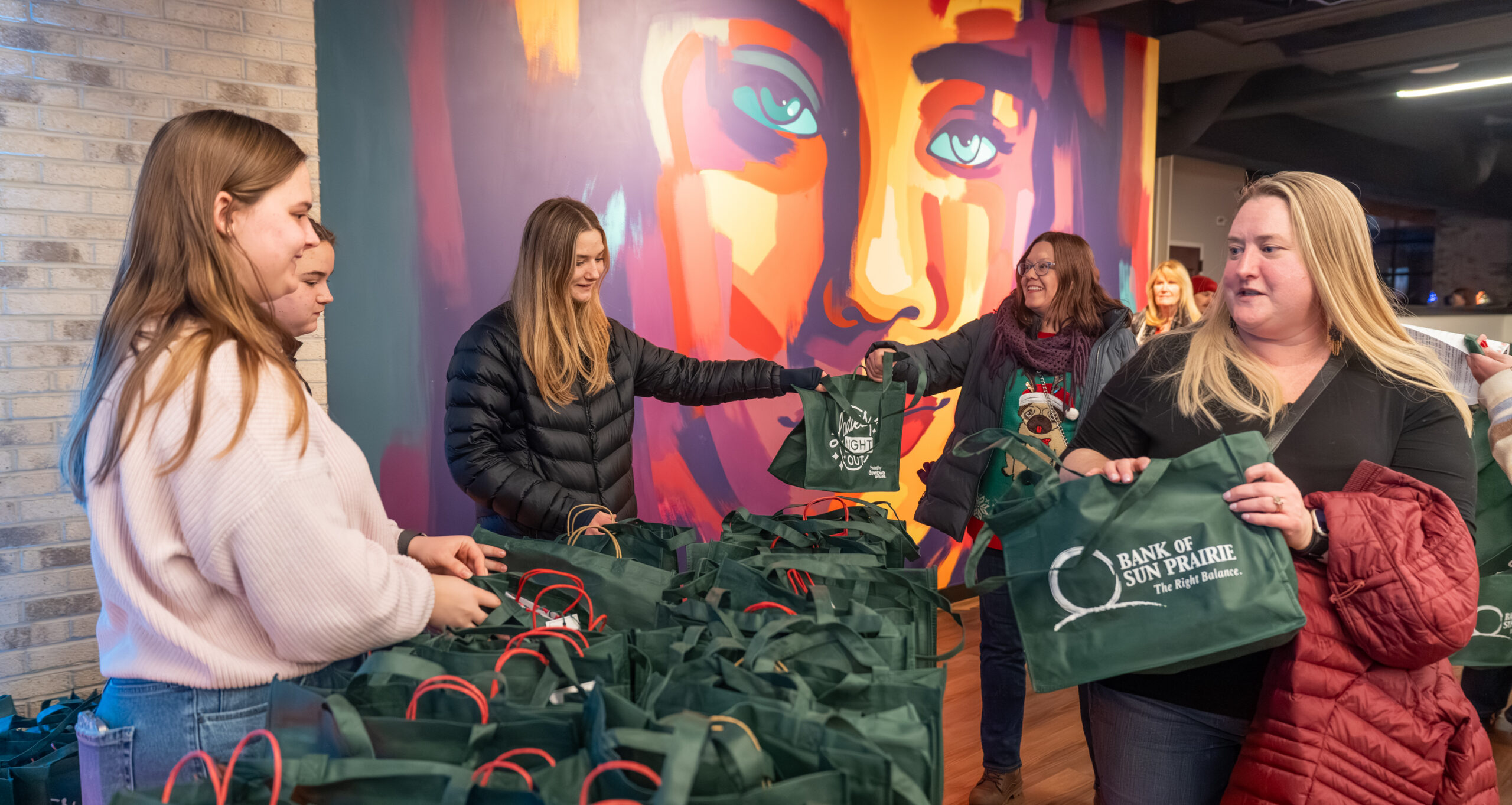 A group of women distributes green tote bags labeled “Bank of Sun Prairie” in a room with a colorful mural in the background.