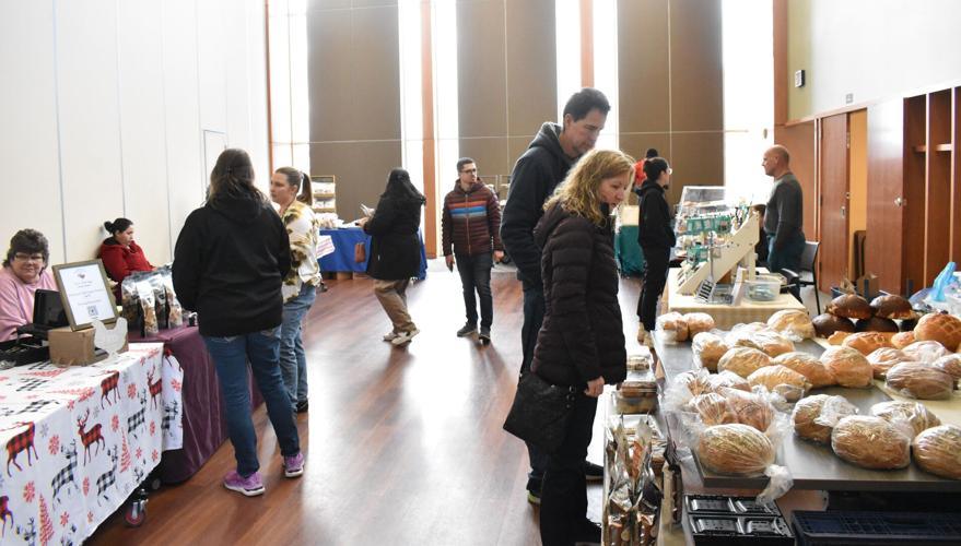 Several people browse tables with baked goods, snacks, and other items at an indoor market with tall windows and wooden floor.