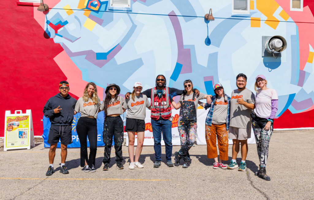 A group of nine people stands in front of a colorful mural, posing for a photo outdoors on a sunny day.