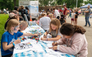Children sit at a table outdoors, drawing on white T-shirts with markers during a community event. Other people are gathered and walking in the background.