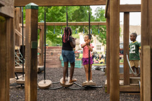 Three children play on a wooden playground structure; two stand on suspended platforms while one observes nearby. Trees and other people are visible in the background.