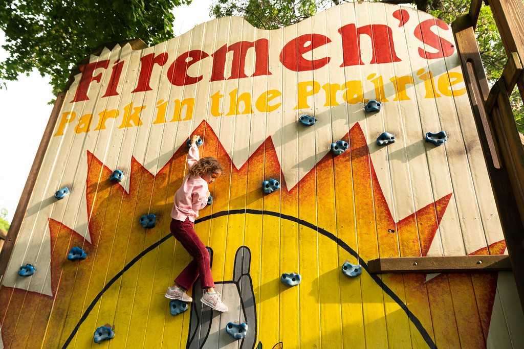 A child climbs a rock wall at Firemen’s Park in the Prairie, with sunlight casting shadows on the painted wooden structure.