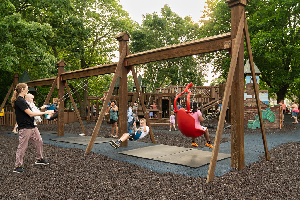 Children and adults enjoy various swings and playground equipment in a wooded outdoor park on a sunny day.