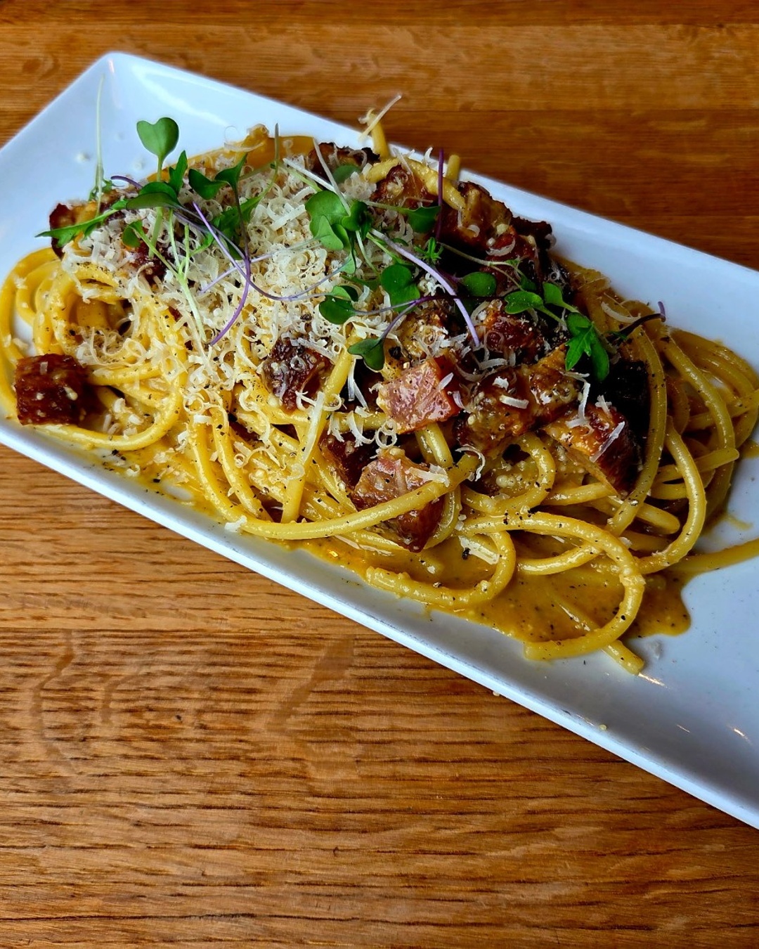 A rectangular white plate with spaghetti pasta topped with grated cheese, herbs, and pieces of meat, set on a wooden table.