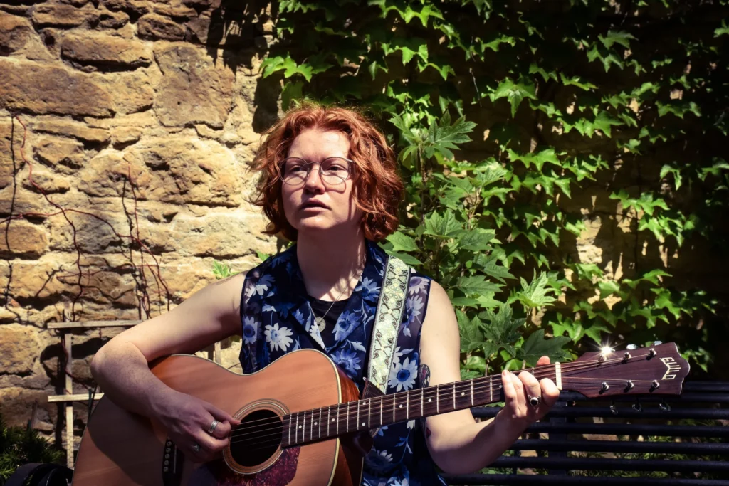 A person with short curly hair and glasses plays an acoustic guitar while sitting outside against a stone wall with green ivy.