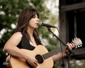 A woman plays an acoustic guitar and sings into a microphone on an outdoor stage, with trees in the background.
