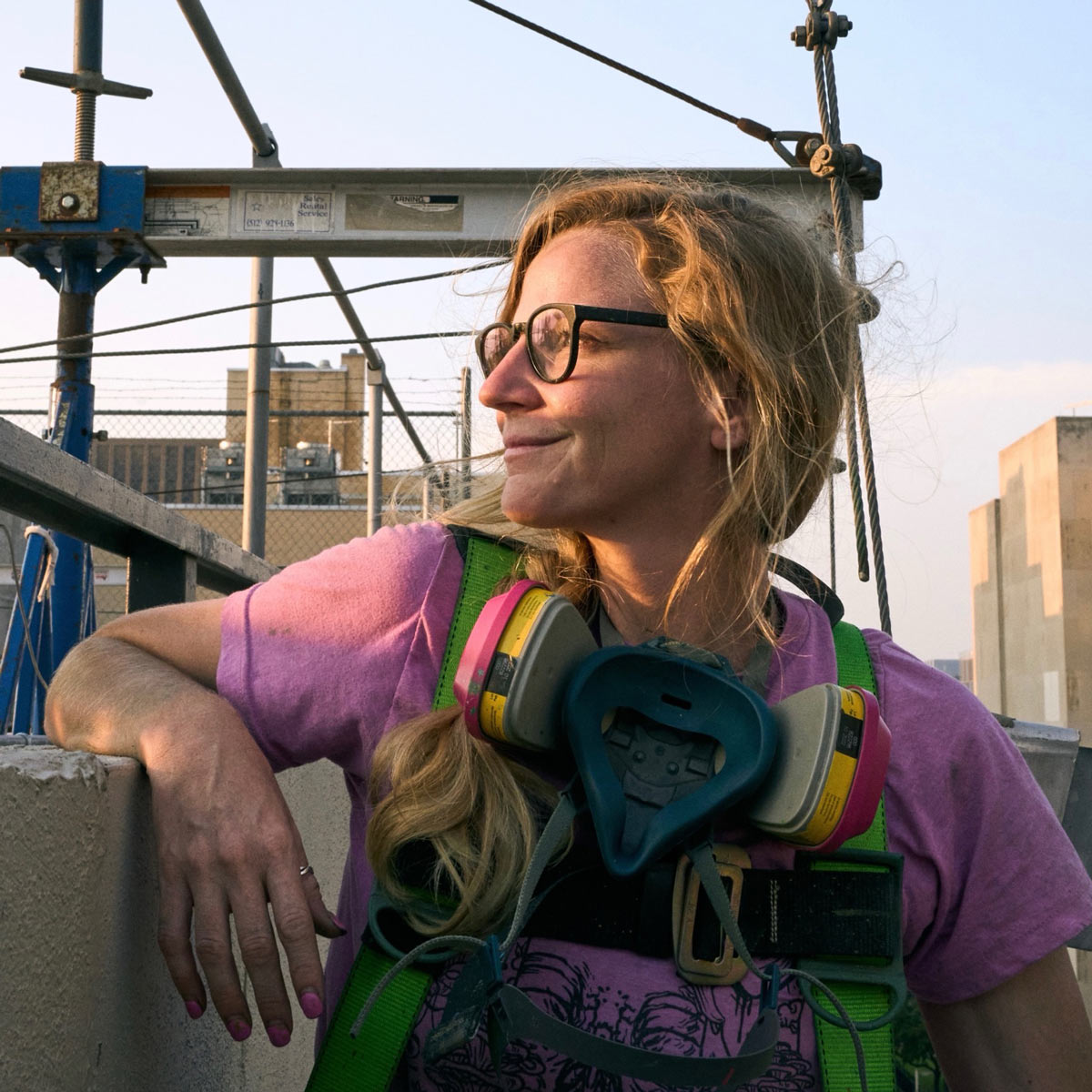 A woman in safety gear and glasses leans on a building ledge at a construction site, with city buildings in the background.