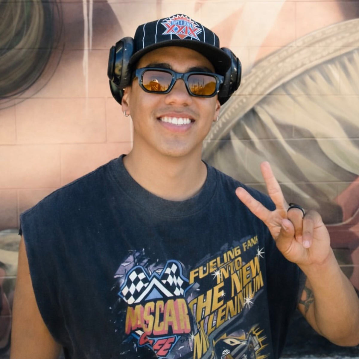 Smiling person wearing sunglasses, headphones, and a graphic t-shirt flashes a peace sign while standing in front of a mural.
