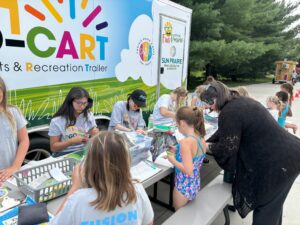 Children and adults participate in arts and crafts activities at an outdoor recreation trailer event, with tables set up beside a colorful trailer.