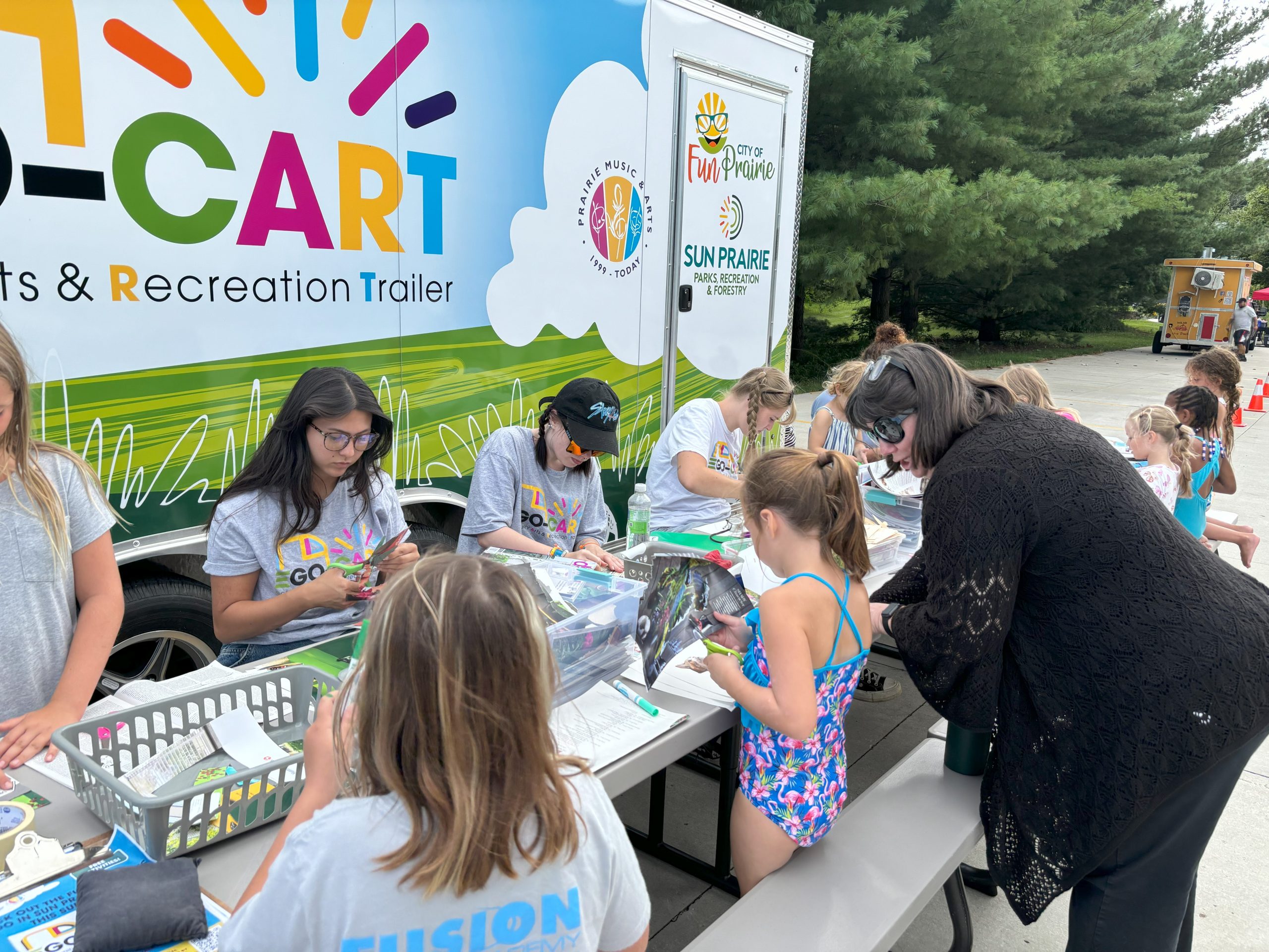 Children and adults participate in arts and crafts activities at an outdoor recreation trailer event, with tables set up beside a colorful trailer.