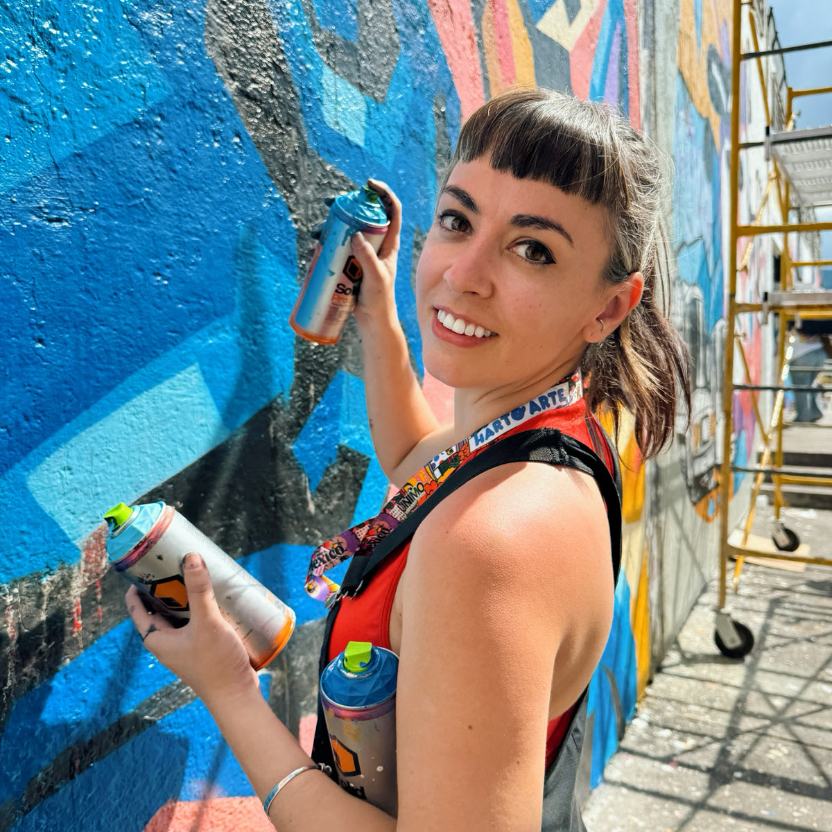 A woman holding spray paint cans paints a colorful mural on an outdoor wall, looking back and smiling at the camera.