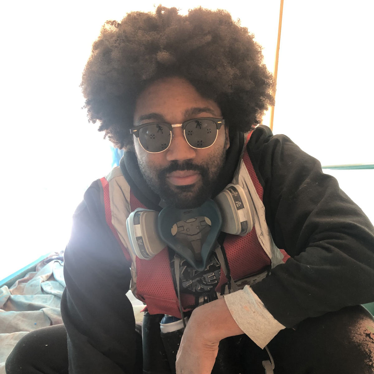 Person with an afro hairstyle wearing sunglasses, a red safety vest, a respirator mask, and a black hoodie, sitting indoors on a tarp-covered floor.