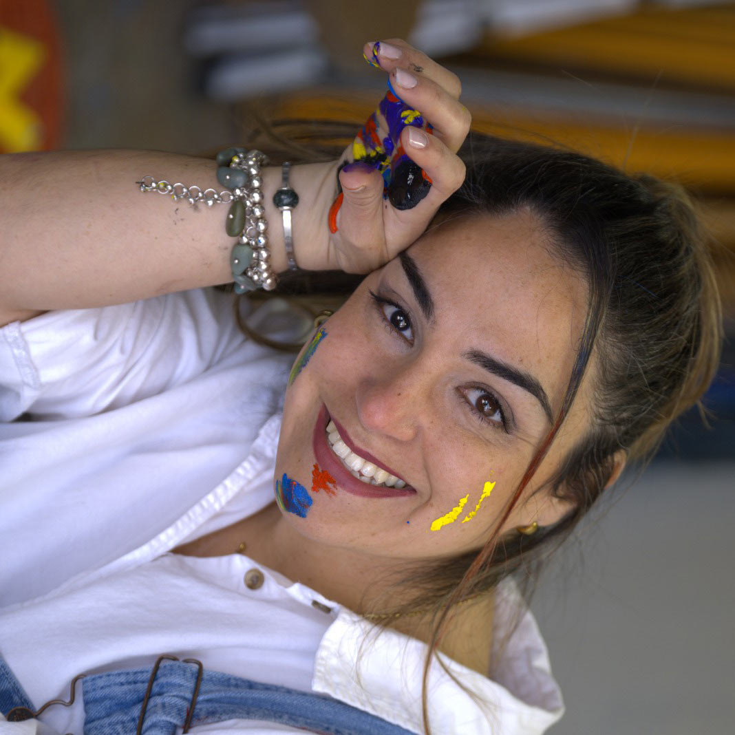 Woman smiling with colorful paint on her face and hand, wearing a white shirt, denim overalls, and bracelets, posing indoors.