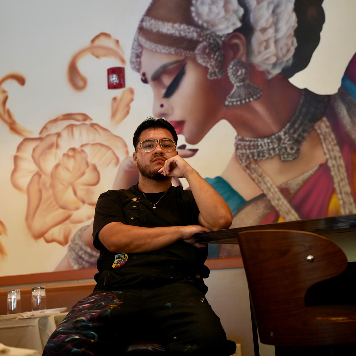 A man sits at a table in front of a large mural depicting a woman in traditional attire and jewelry, with flowers in the background.